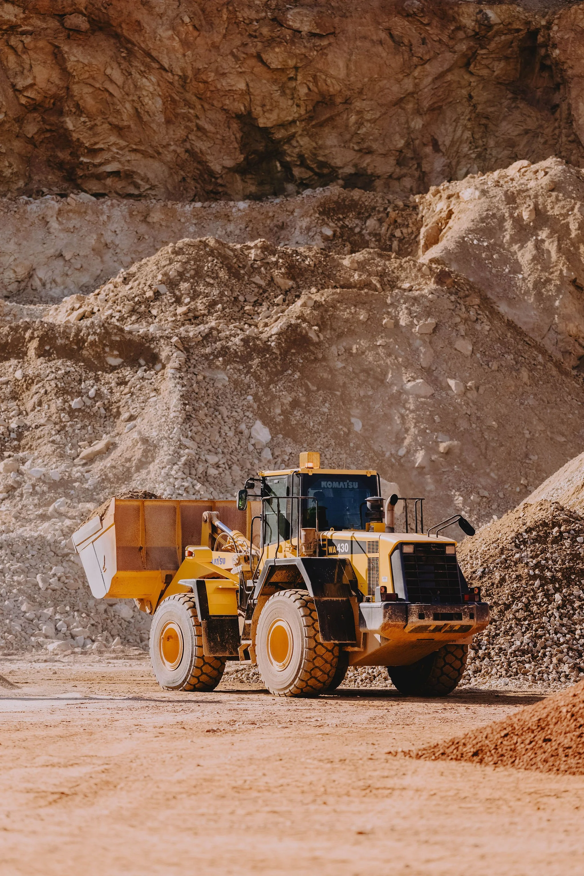 A yellow Komatsu WA430 wheel loader operating on a construction site with large piles of dirt and rocky terrain in the background.