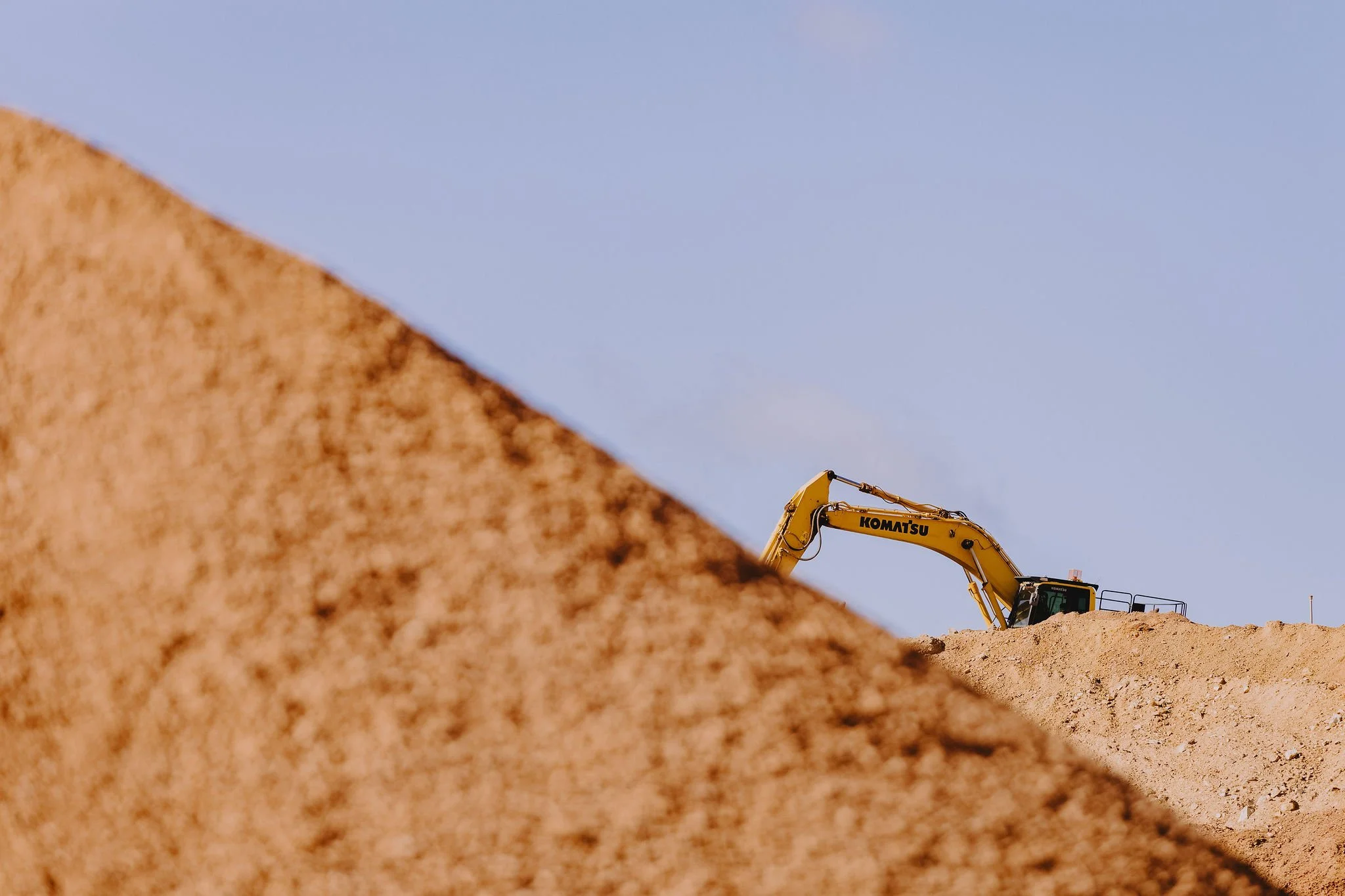 A yellow Komatsu excavator on a mound of dirt at a construction site, with a clear blue sky in the background.