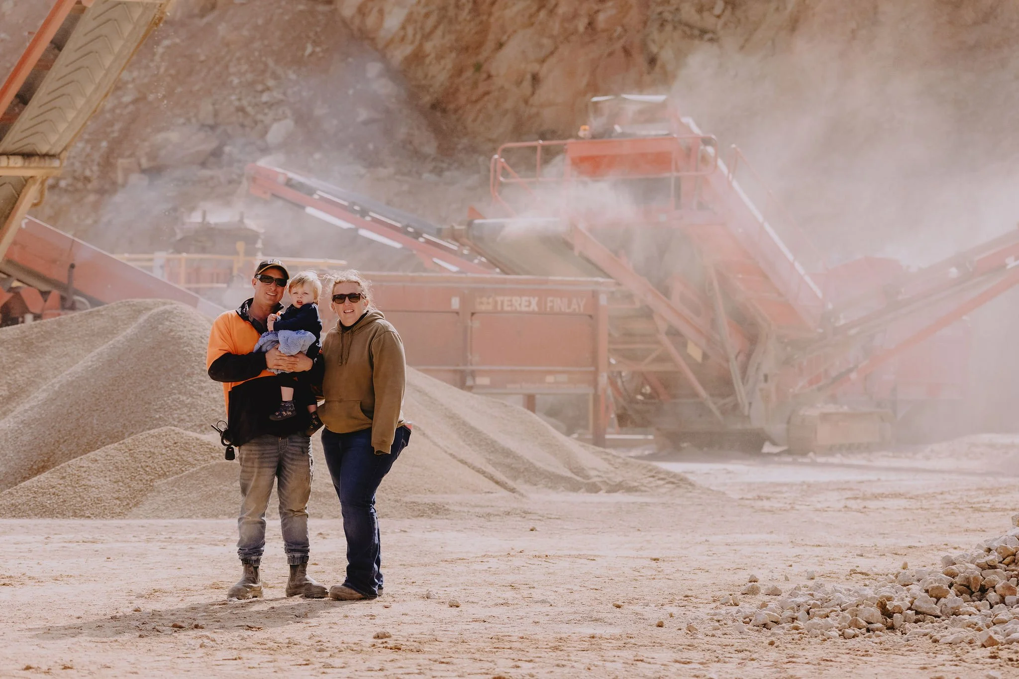 Three people, two adults and a child, standing on a mound of dirt near a construction or mining site with large machinery and dust in the background.