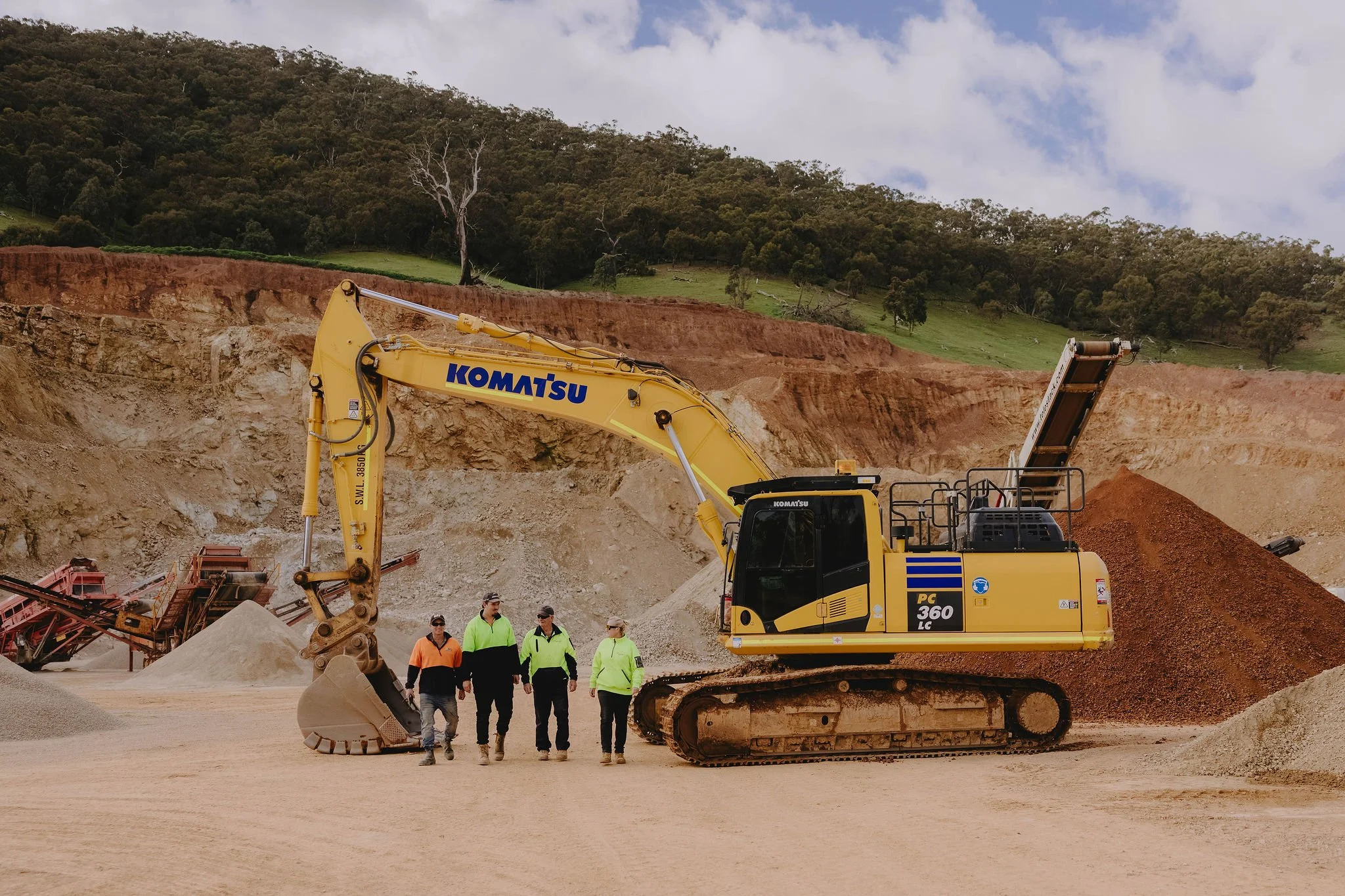 A yellow Komatsu excavator working on a construction site with a group of four people and green hills in the background.