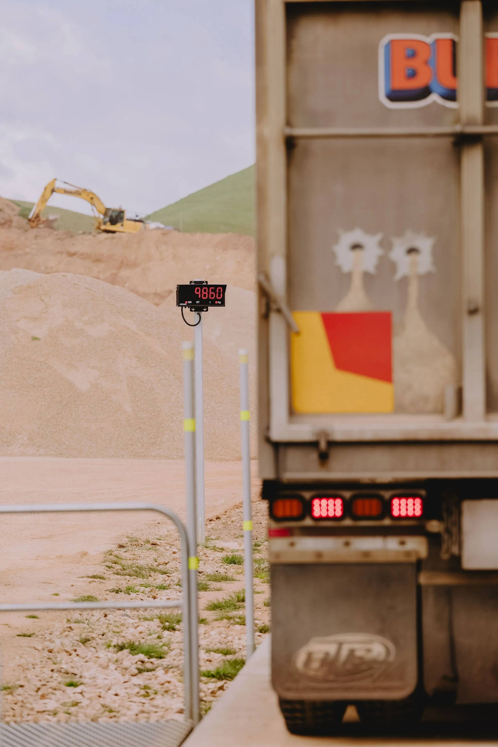 A truck at a border crossing, with a digital weigh station display showing 9860 pounds, and construction equipment on a hillside in the background.