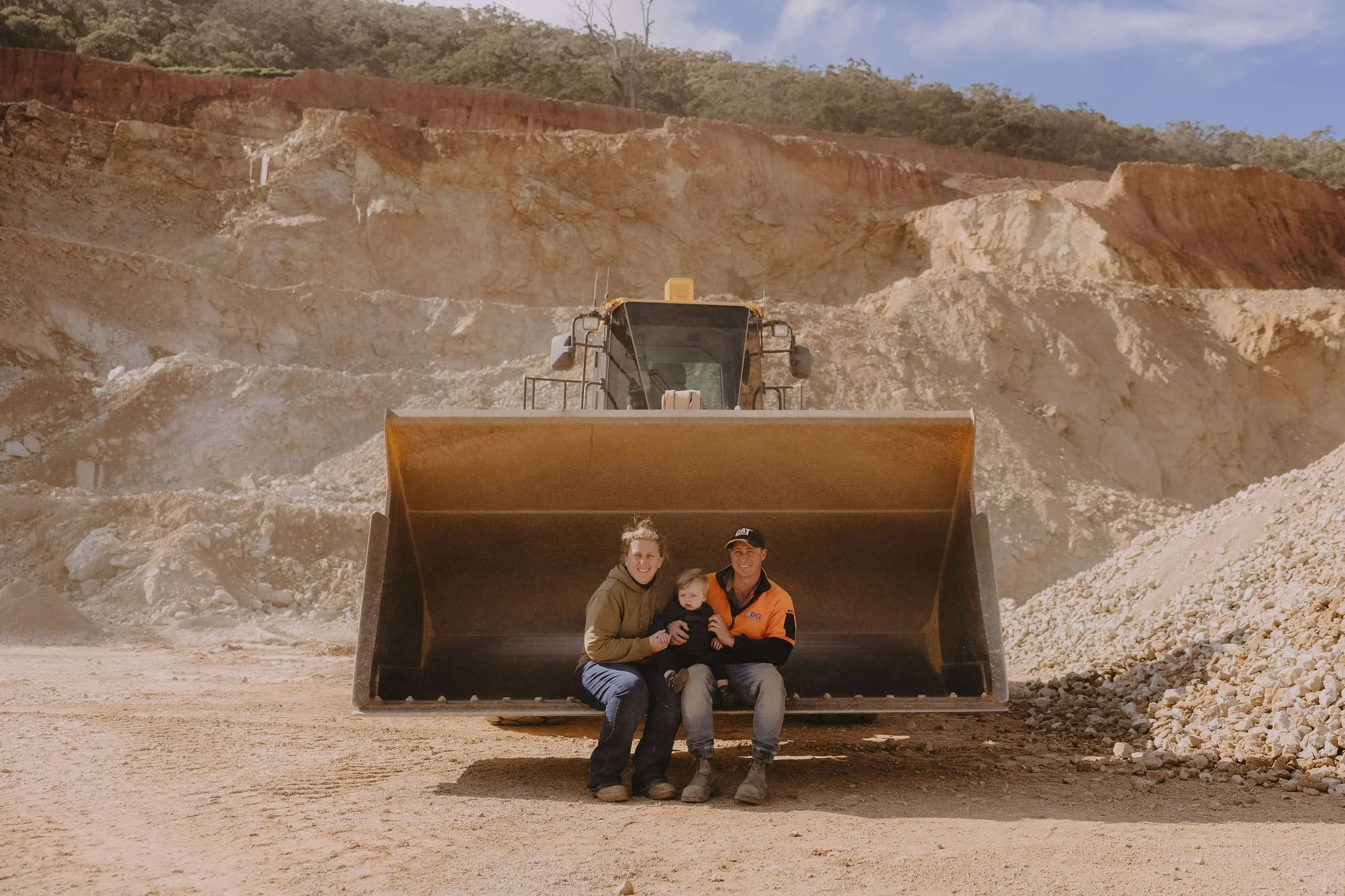 Three people sitting inside the bucket of a large yellow construction vehicle, smiling, with a woman holding a small child, and a man in an orange jacket. Behind them is a rugged, rocky landscape with large dirt and rock piles and a bulldozer or similar heavy machinery.