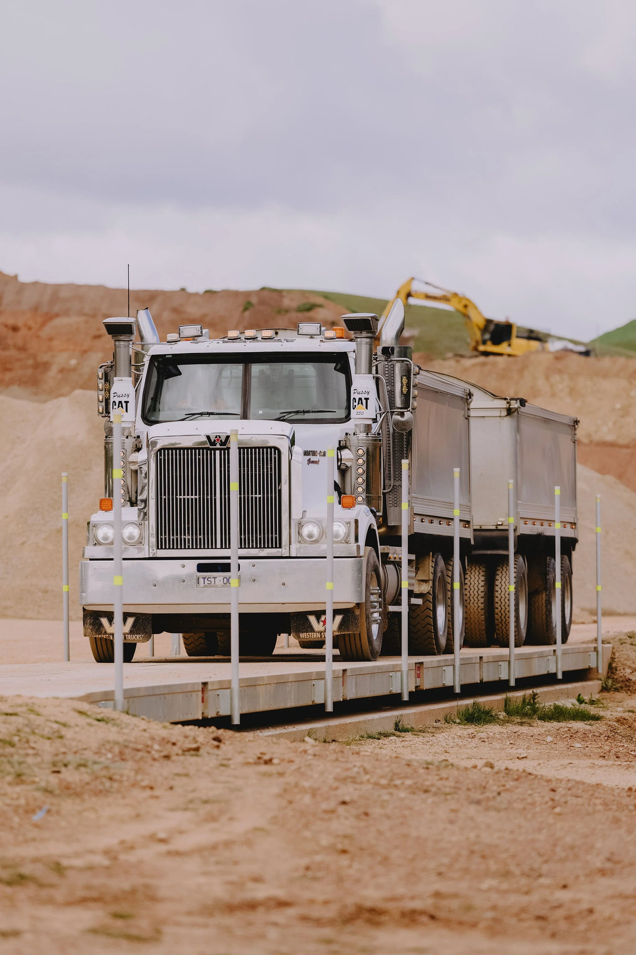 A large white semi-truck with a flatbed trailer carrying construction equipment on a dirt road with a background of hills and overcast sky.