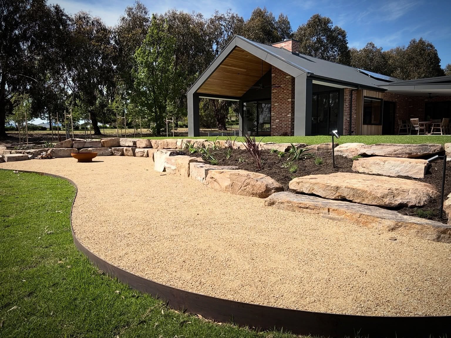 A landscaped backyard featuring a curved gravel pathway with a border of green grass, a flower bed with large rocks and plants, a small clay pot, and a modern house with a covered patio and wooden dining table in the background. Tall trees surround t