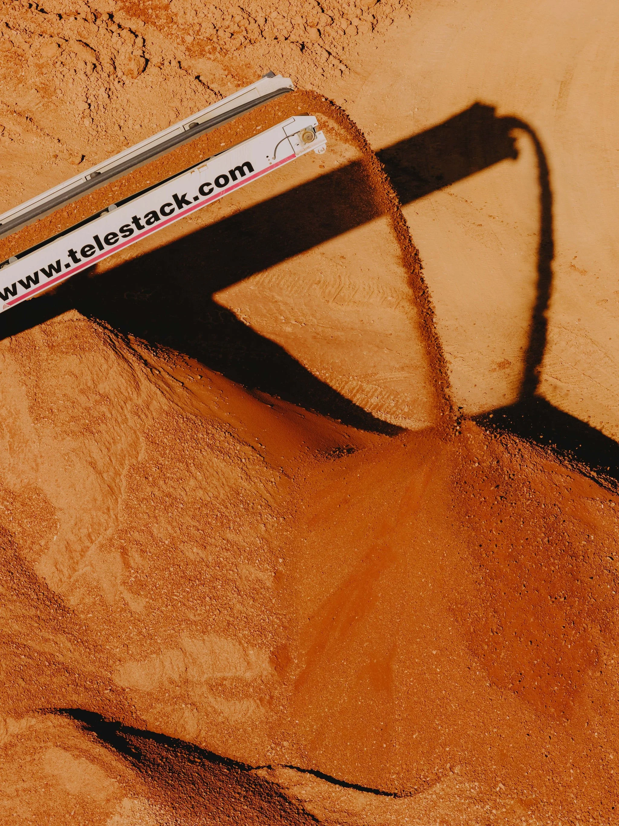 Close-up of a survey marker on a dirt ground, with a shadow cast on the ground showing part of the marker and a shadow of a person or object nearby.