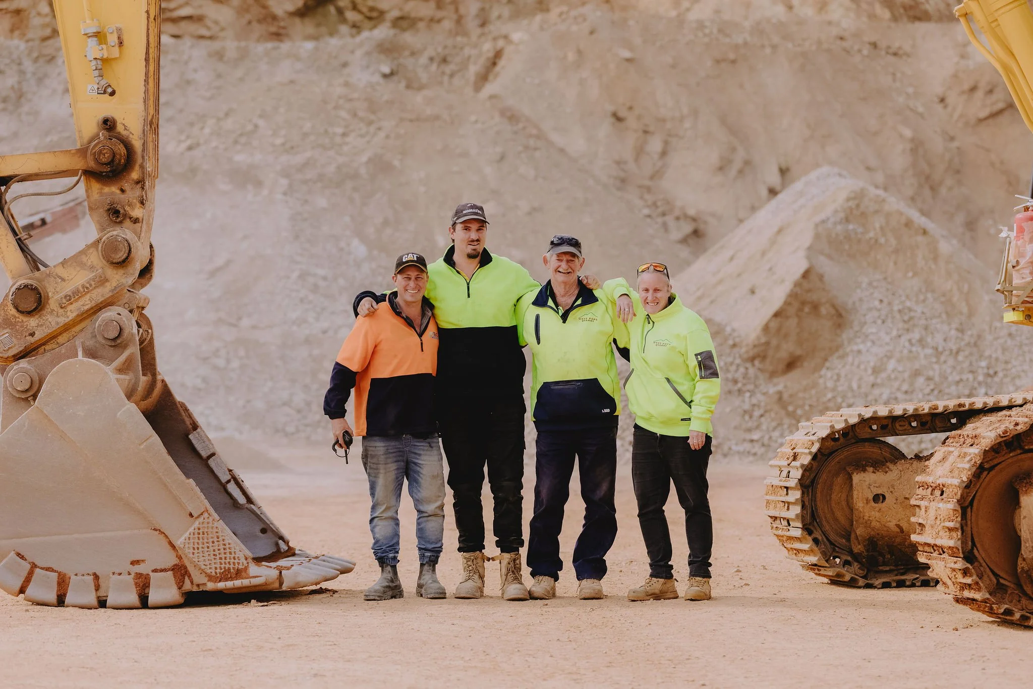 Four smiling men standing together outdoors behind a large construction vehicle, with a dirt hill in the background, wearing safety jackets and hats.