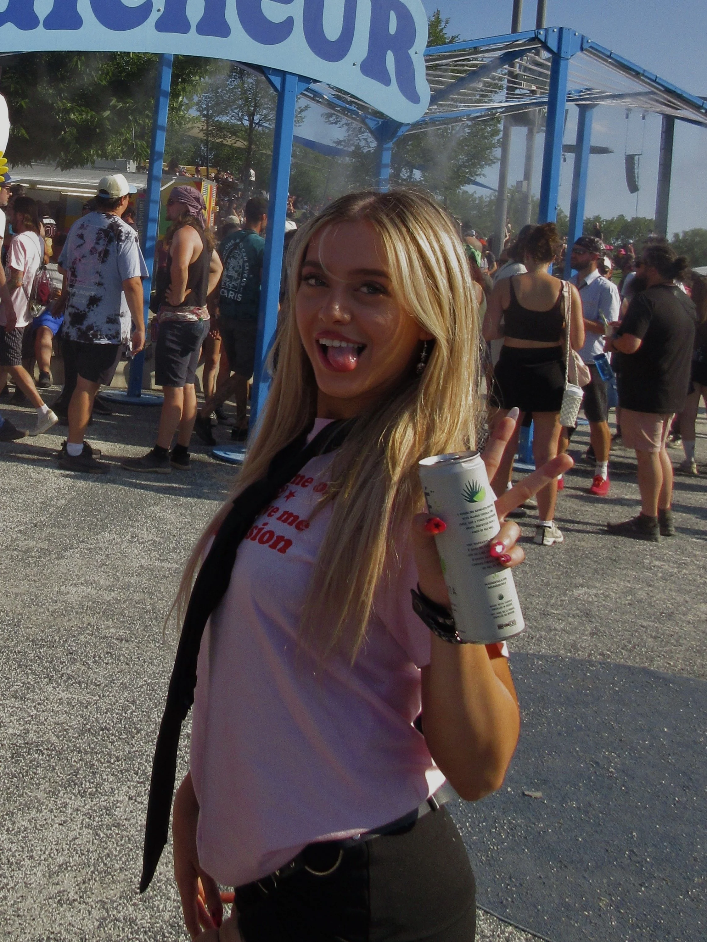 A young woman with blonde hair making a face with her tongue out, holding a white can with a green and yellow logo in her right hand, at an outdoor event with many people in the background.