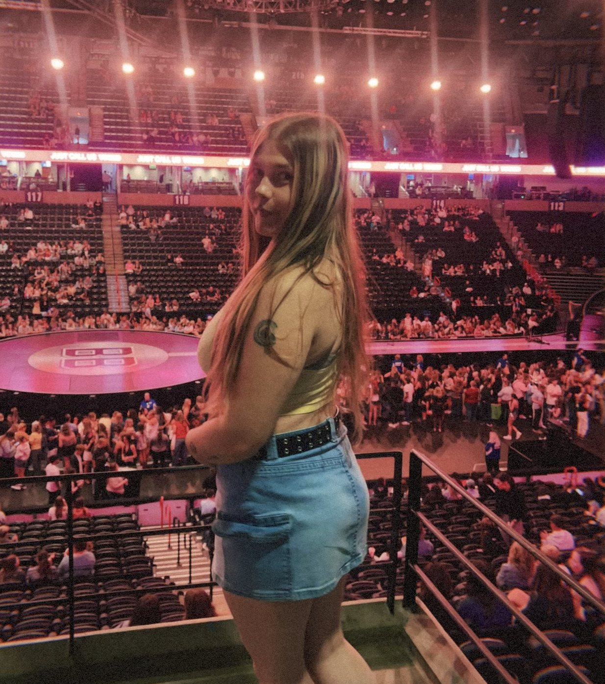 A woman with long hair, wearing a yellow top and denim shorts, is standing in an arena with a wrestling ring in the background. The arena is filled with spectators, and the lighting has a pink hue.