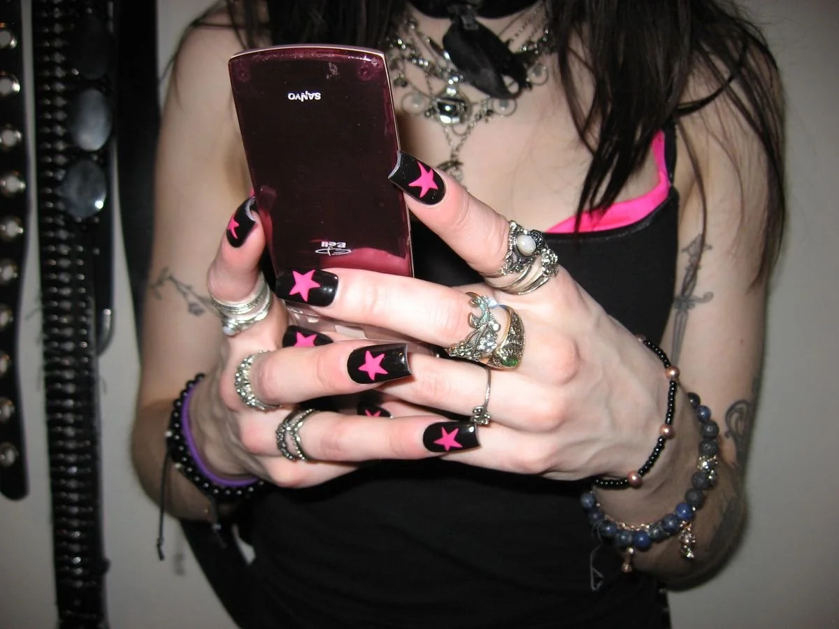 A woman with dark hair, wearing a black tank top with pink accents, is holding a pink and black flip phone. She has multiple rings, bracelets, and decorated nails with black polish and pink stars.