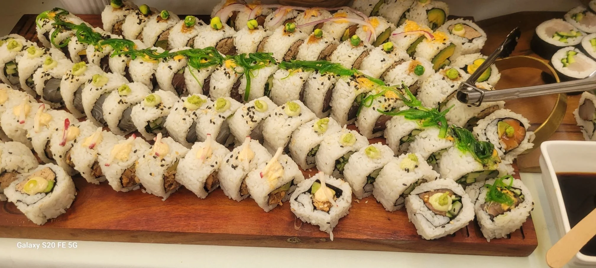 Assorted sushi rolls on a wooden platter, garnished with green onions, seaweed, and sesame seeds. There are soy sauce containers nearby.