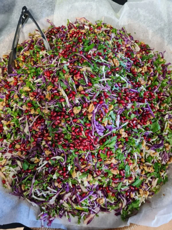 A large colorful chopped vegetable salad with purple cabbage, chopped herbs, pomegranate seeds, and walnuts, placed on parchment paper with tongs on the side.