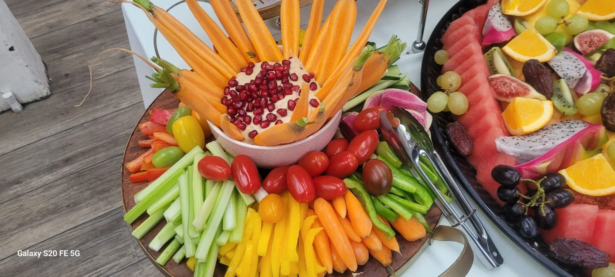 Assorted fresh fruits and vegetables arranged on a platter, including carrots, cherry tomatoes, celery, yellow bell peppers, and a bowl of dip garnished with pomegranate seeds, with additional sliced fruits like watermelons, dragon fruit, oranges, gr
