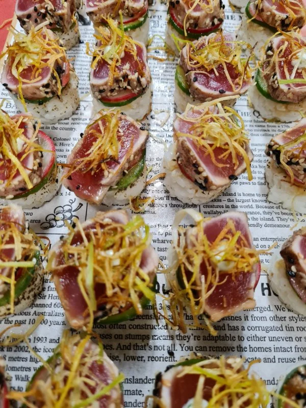 Close-up of assorted sushi bites topped with slices of raw tuna, thinly sliced vegetables, and shredded garnish, arranged on a newspaper background.