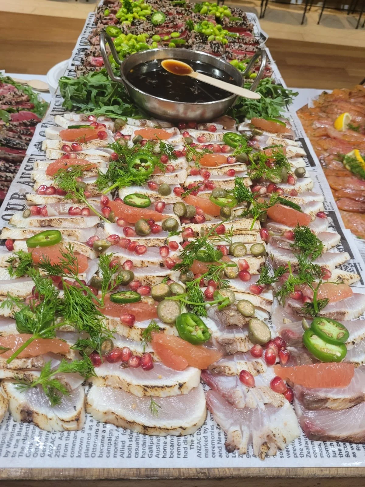A platter of sliced raw fish garnished with herbs, sliced jalapeños, pomegranate seeds, and capers, with a bowl of dark dipping sauce in the center, on a table covered with newspaper.