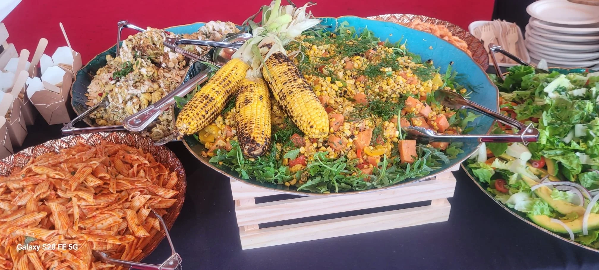A large serving platter of grilled corn on the cob and mixed vegetable salad, surrounded by plates of pasta, salad, and small takeout containers at a buffet table.