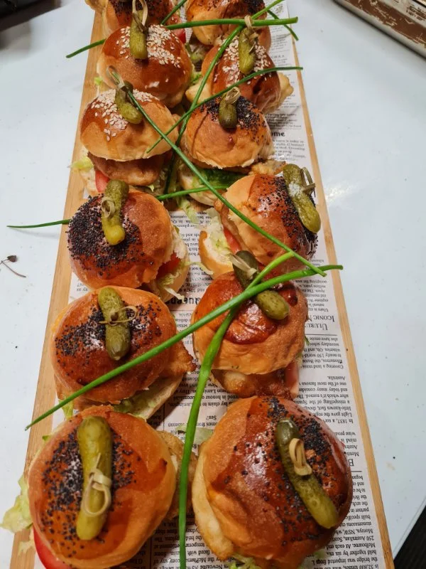 A wooden platter with small sandwiches topped with pickles, poppy seeds, and garnished with green chives, arranged on a sheet of paper.