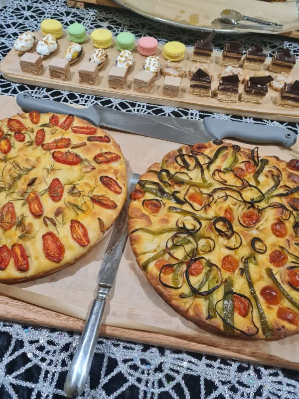 Two pizzas on a wooden serving board, one with cherry tomatoes and rosemary, the other with sliced green peppers, cherry tomatoes, and sliced black peppers. Behind them, there is a knife and a large fork. In the background, a table with a lace tablec