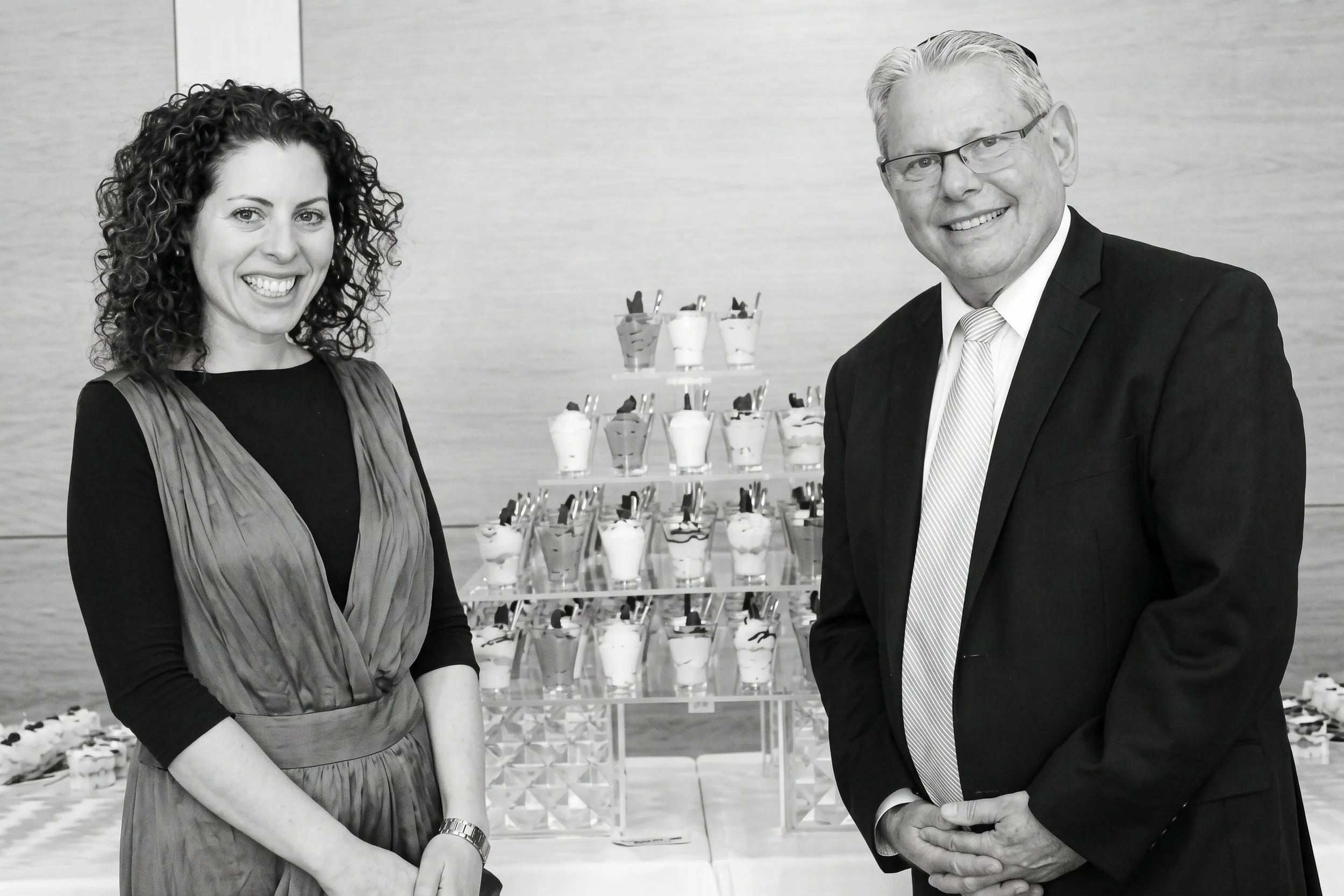 A woman and a man dressed in formal attire smiling at the camera at a dessert reception, with a display of small desserts in the background.