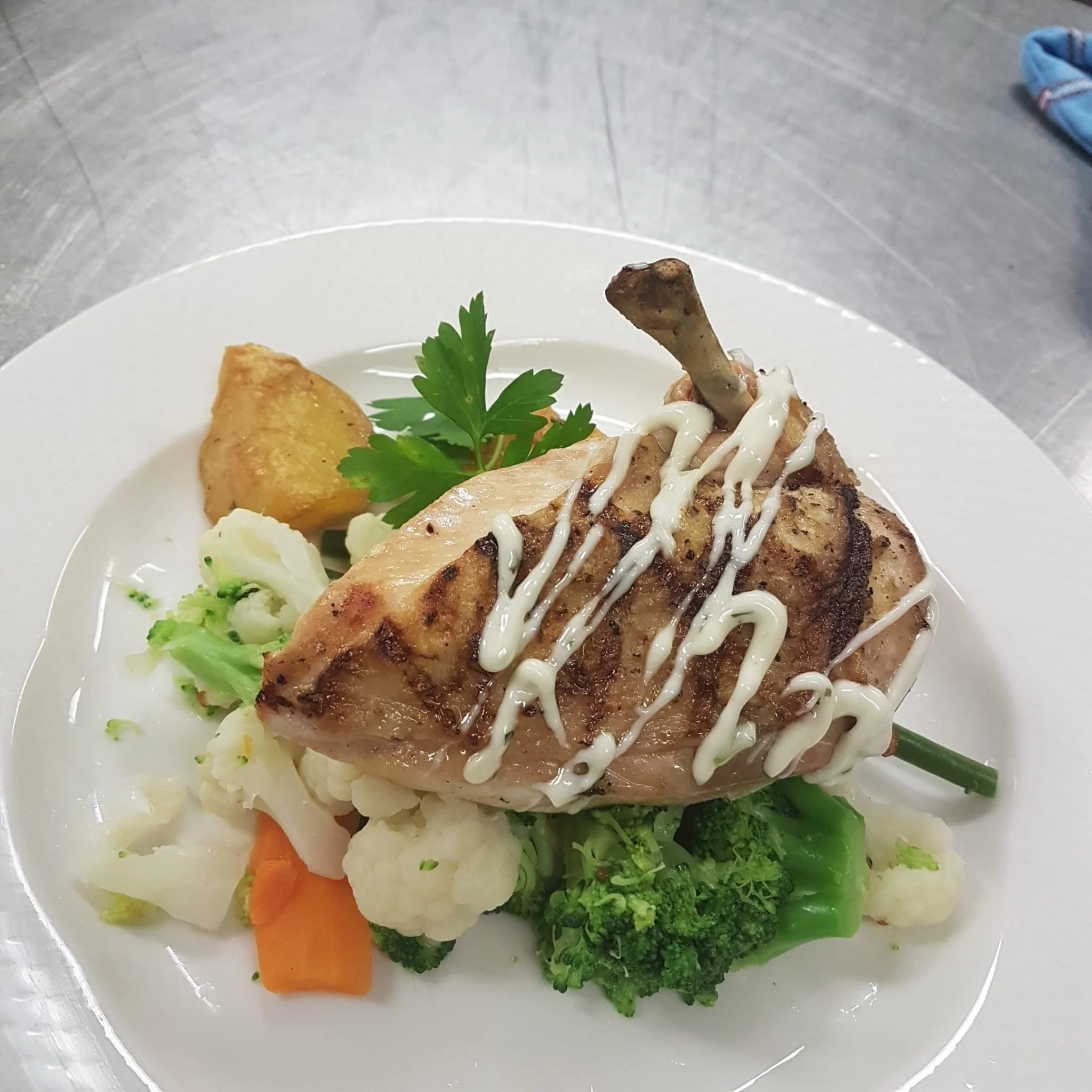 A plate with grilled chicken topped with mayonnaise sauce, garnished with parsley, and served with roasted potatoes, carrots, broccoli, and lettuce.