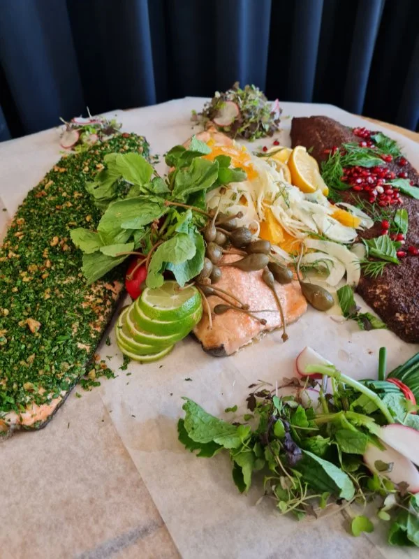 Assorted smoked and cured fish with herbs, radishes, sliced lime, and lemon slices, arranged on parchment paper for serving.