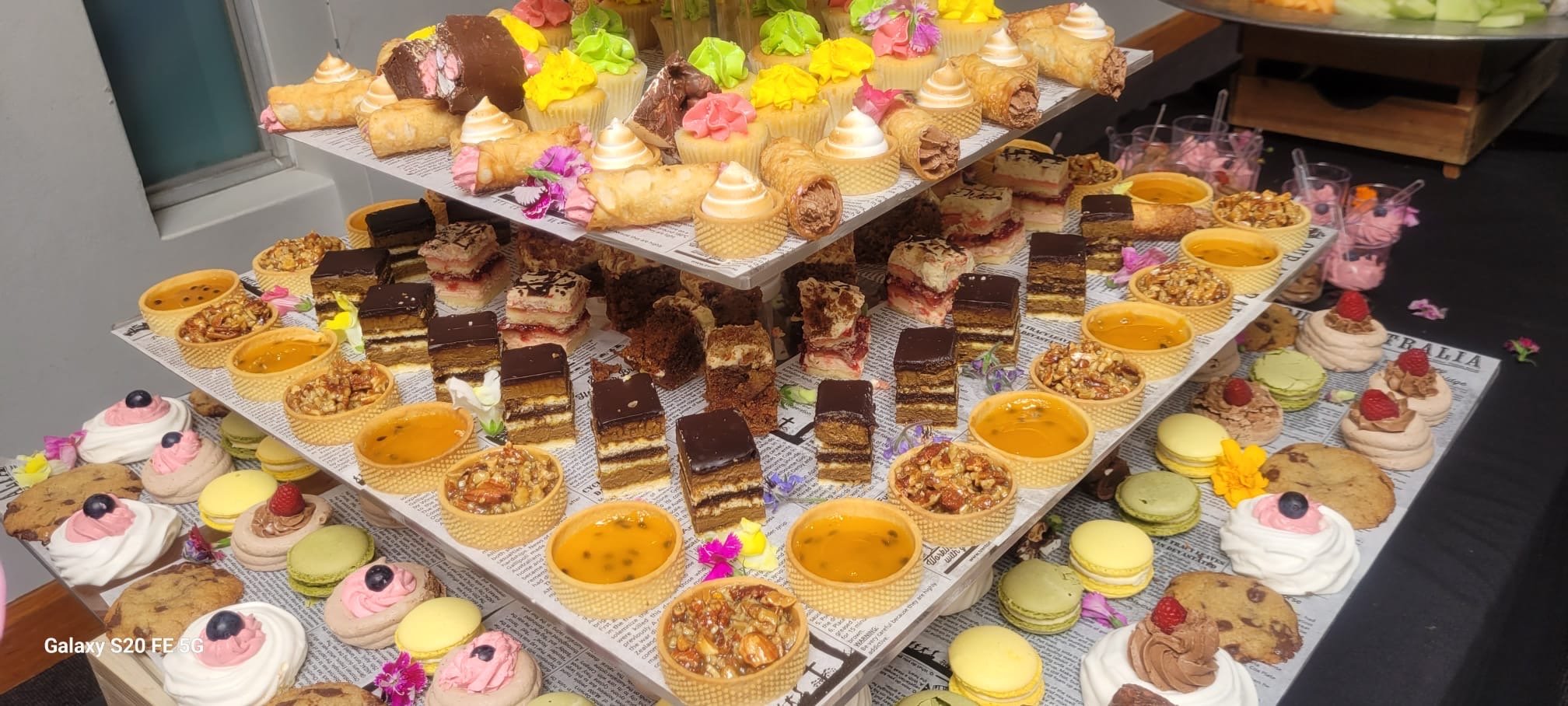 A display of colorful assorted desserts on three-tiered trays, including mini cupcakes with pink, yellow, and white frosting, pastry rolls, chocolate layered cakes, small puddings in yellow cups, and decorated cookies with berries and flowers.