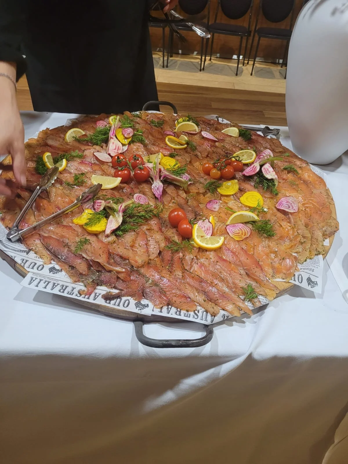 A large platter of sliced raw salmon garnished with cherry tomatoes, lemon wedges, and assorted herbs and vegetables, served on a table.