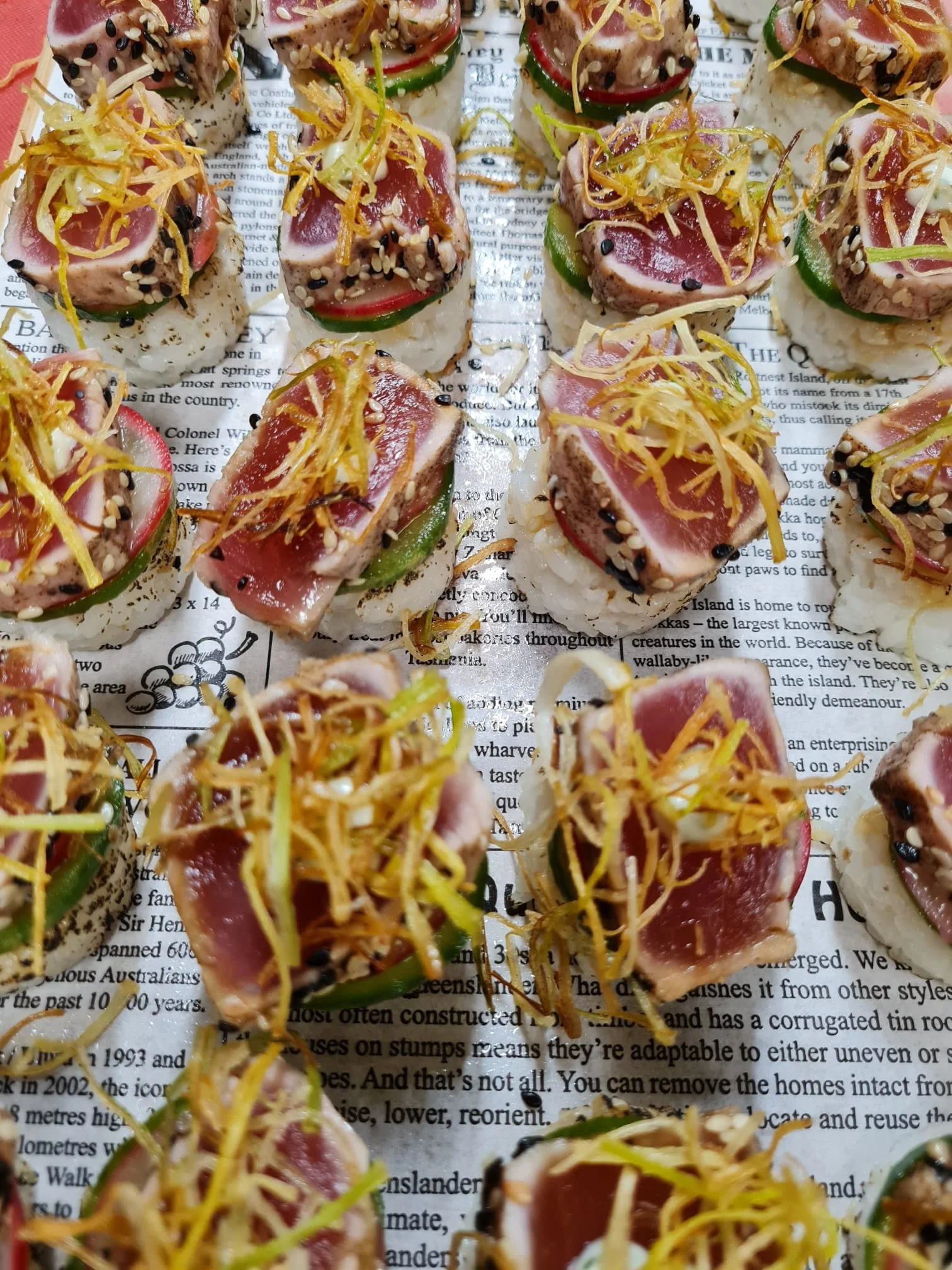 Close-up of sushi bites topped with fresh tuna, sliced vegetables, shredded ginger, and sprinkled sesame seeds on a newspaper background.