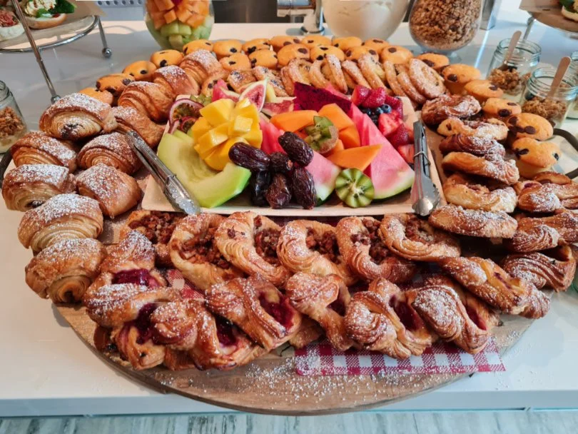Assorted pastries including croissants and Danish pastries surrounding a tray of sliced fresh fruit, with strawberries, kiwi, watermelon, cantaloupe, and grapes, on a table for a buffet or brunch setting.