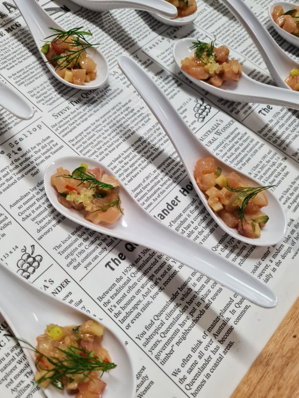 Cups of raw diced fish with herbs and garnish on white ceramic spoons, placed on a newspaper-covered surface.