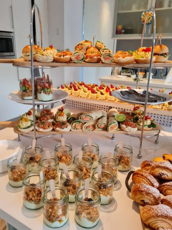 A three-tiered serving tray with assorted sandwiches, wraps, and small desserts. The bottom tier has jars of yogurt with granola and spoons. The middle tier features mini sandwiches with various fillings and lime wedges. The top tier displays more sa