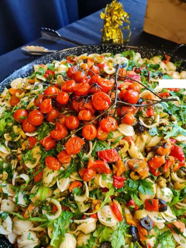Colorful pasta salad with cherry tomatoes, bell peppers, olives, and fresh herbs on a large black platter, with a yellow flower and utensils nearby.