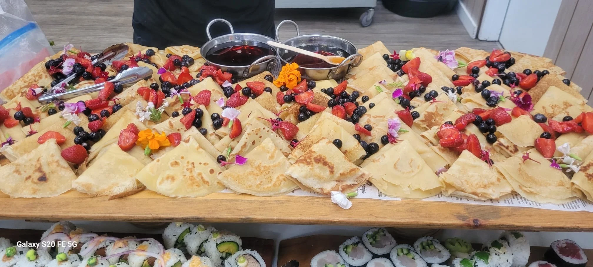 A table of crepes topped with strawberries, blueberries, whipped cream, and edible flowers, with small containers of syrup in the center.
