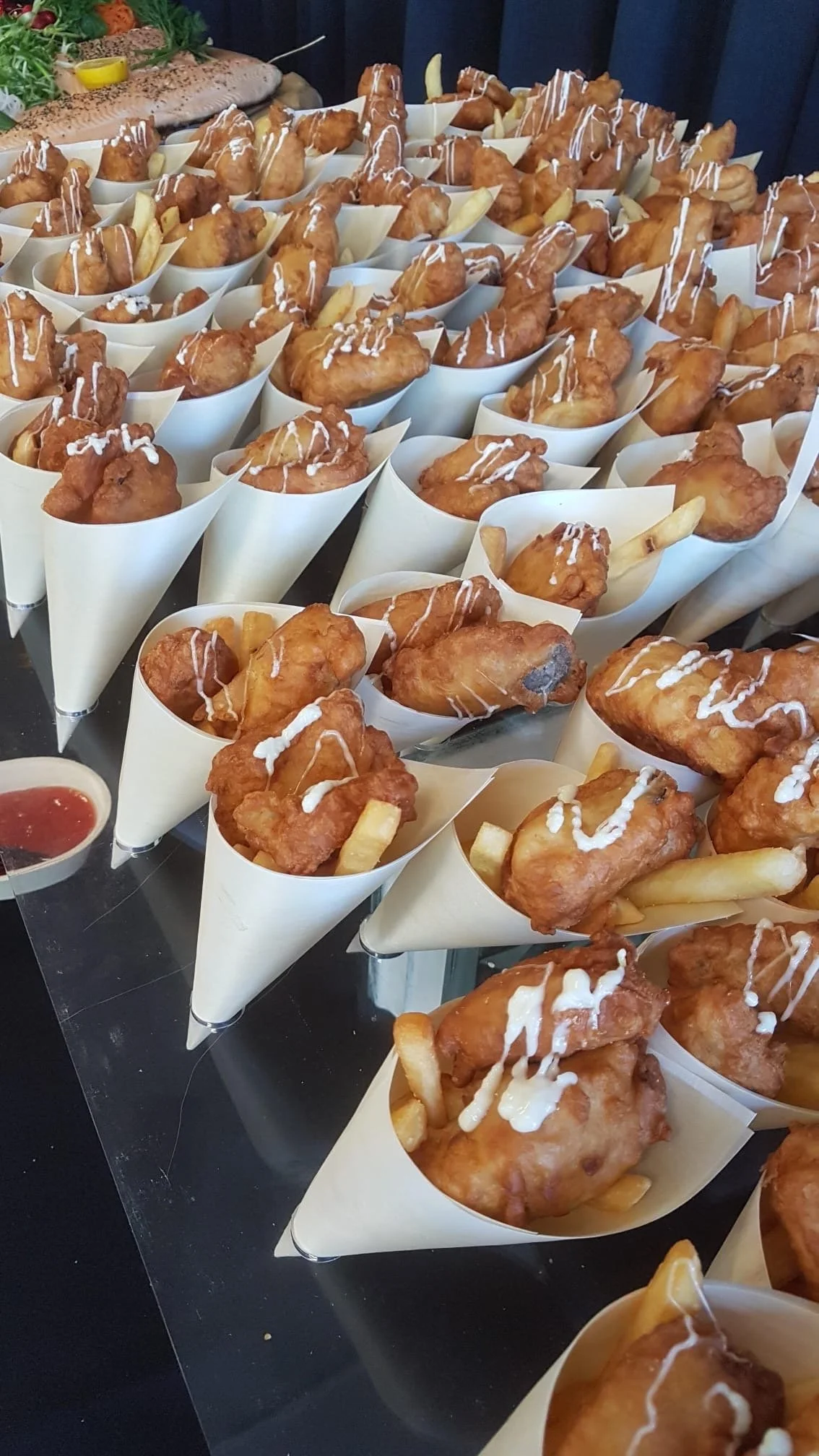 Multiple servings of fried chicken drumsticks with French fries topped with white sauce, served in white paper cones on a black table, with a bowl of ketchup on the side.
