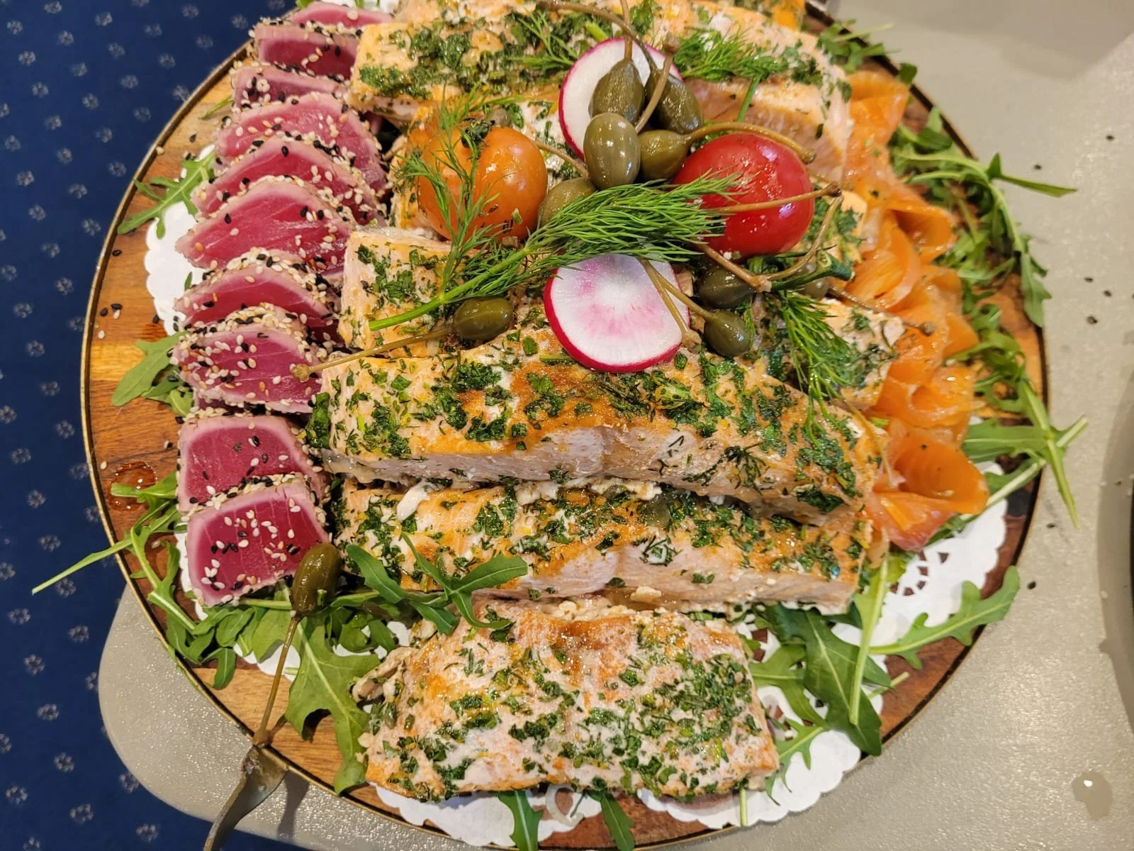 A round wooden platter with slices of seared tuna coated in herbs, topped with fresh dill, radish slices, cherry tomatoes, caper berries, and arugula, garnished with sesame seeds and herbs.