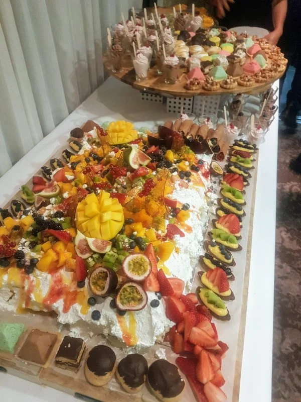 Large dessert table filled with various colorful cakes, cupcakes, fruit, and pastries, with a focus on fruit-topped desserts and decorated sweets.