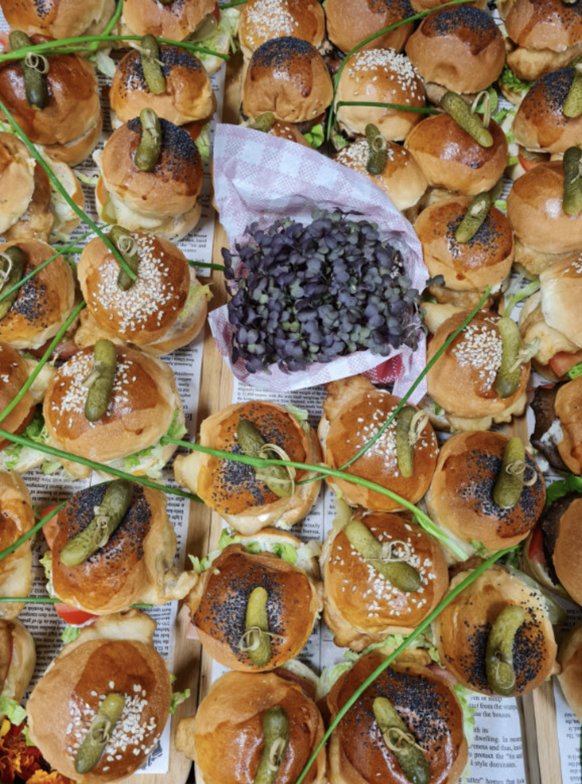 A large tray of mini sandwiches filled with meat, lettuce, and tomato, topped with sesame or poppy seeds and garnished with small pickles and green chives.