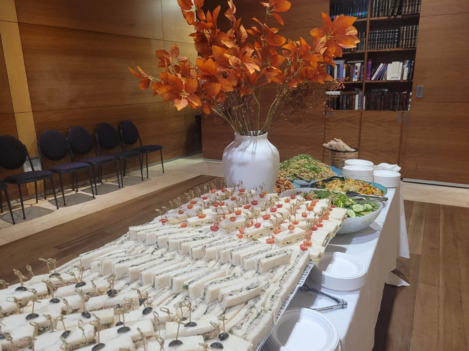 A buffet table with various finger foods, including sandwiches with toothpicks, salads, and sliced vegetables, set up in a room with wood-paneled walls and black chairs.