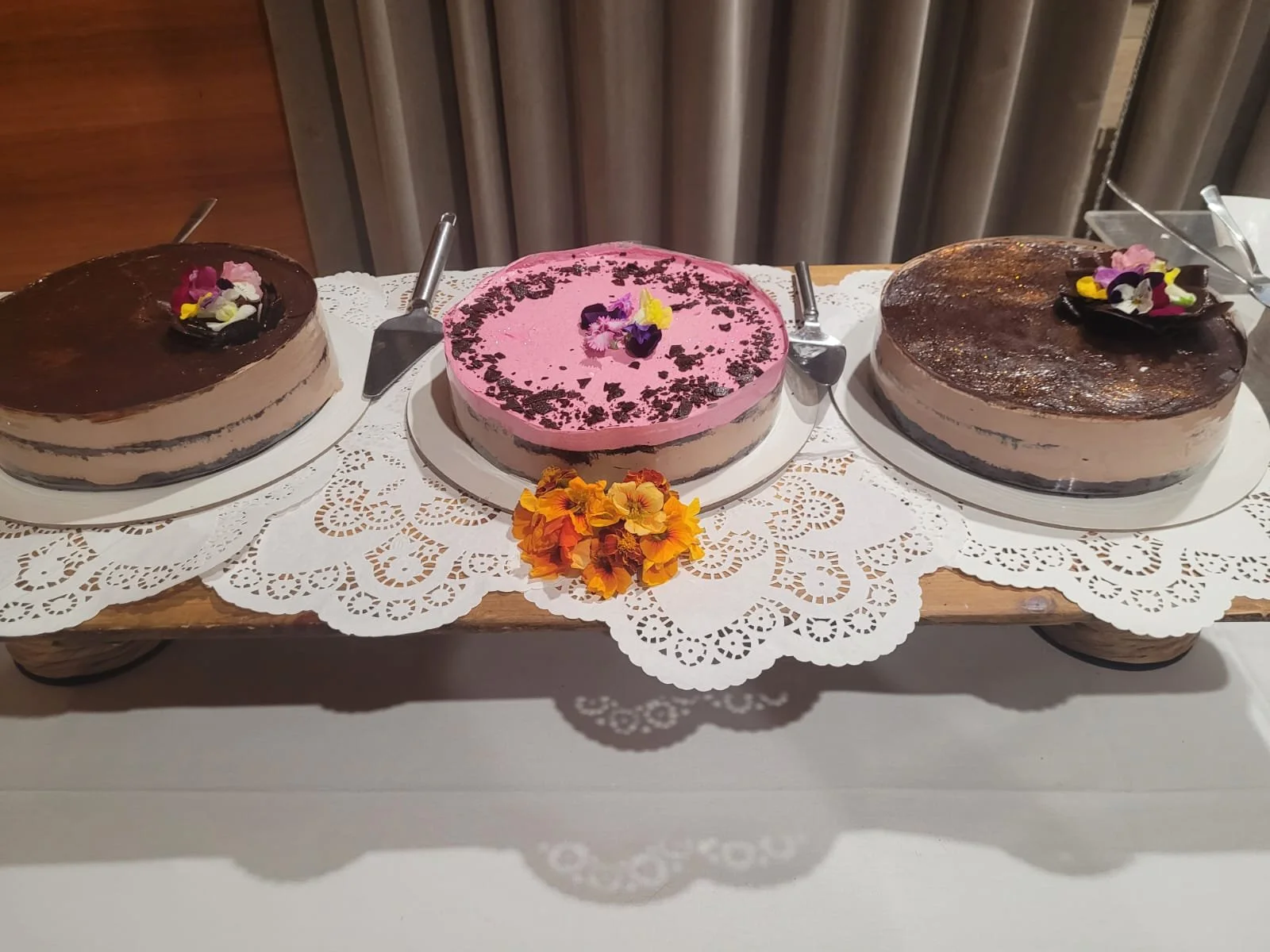 Three decorated cakes on a wooden tray with a lace doily, surrounded by flowers and cake servers.