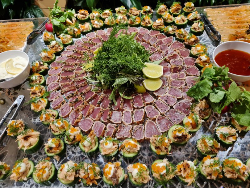 Platter of raw tuna sashimi garnished with lemon wedges in the center, surrounded by cucumber slices topped with seafood and herbs, with dipping sauces on the side.