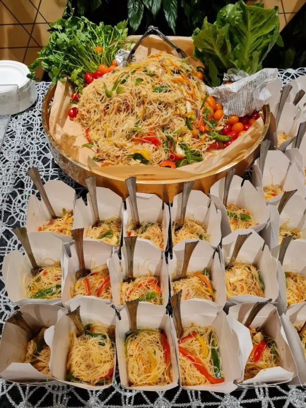 Tray of colorful vegetable noodles garnished with lettuce, cherry tomatoes, and greens, with individual servings of vegetable noodle salad in paper cups arranged around it on a lace tablecloth.