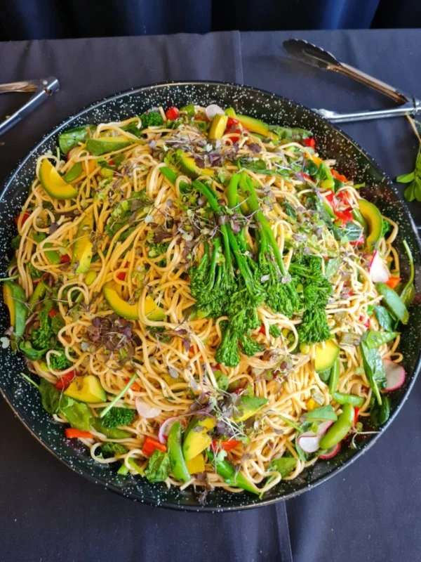 Large black speckled skillet filled with stir-fried noodles and colorful vegetables including broccoli, zucchini, radishes, and bell peppers, garnished with microgreens, on a black table with tongs and a serving spoon.