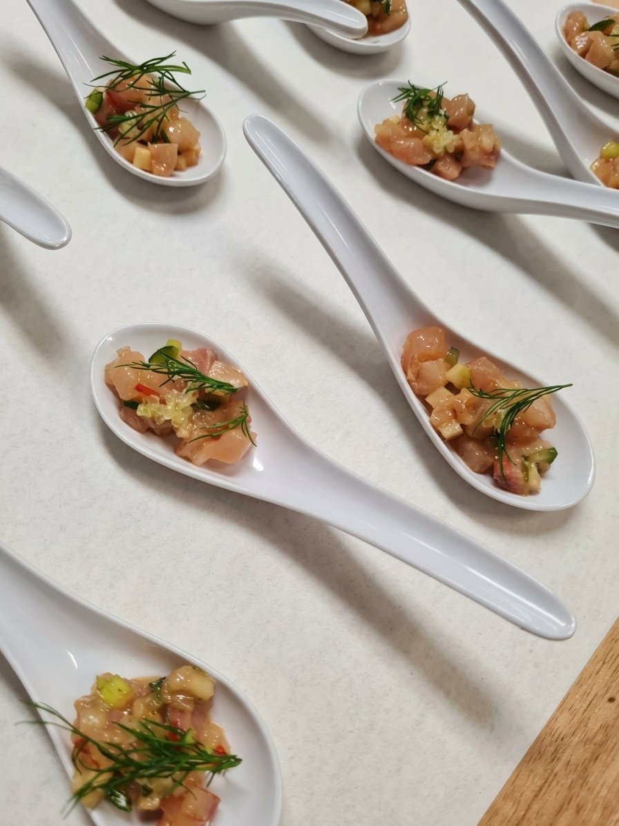 Small white spoons filled with raw diced fish, garnished with herbs and chopped vegetables, arranged on a white surface.