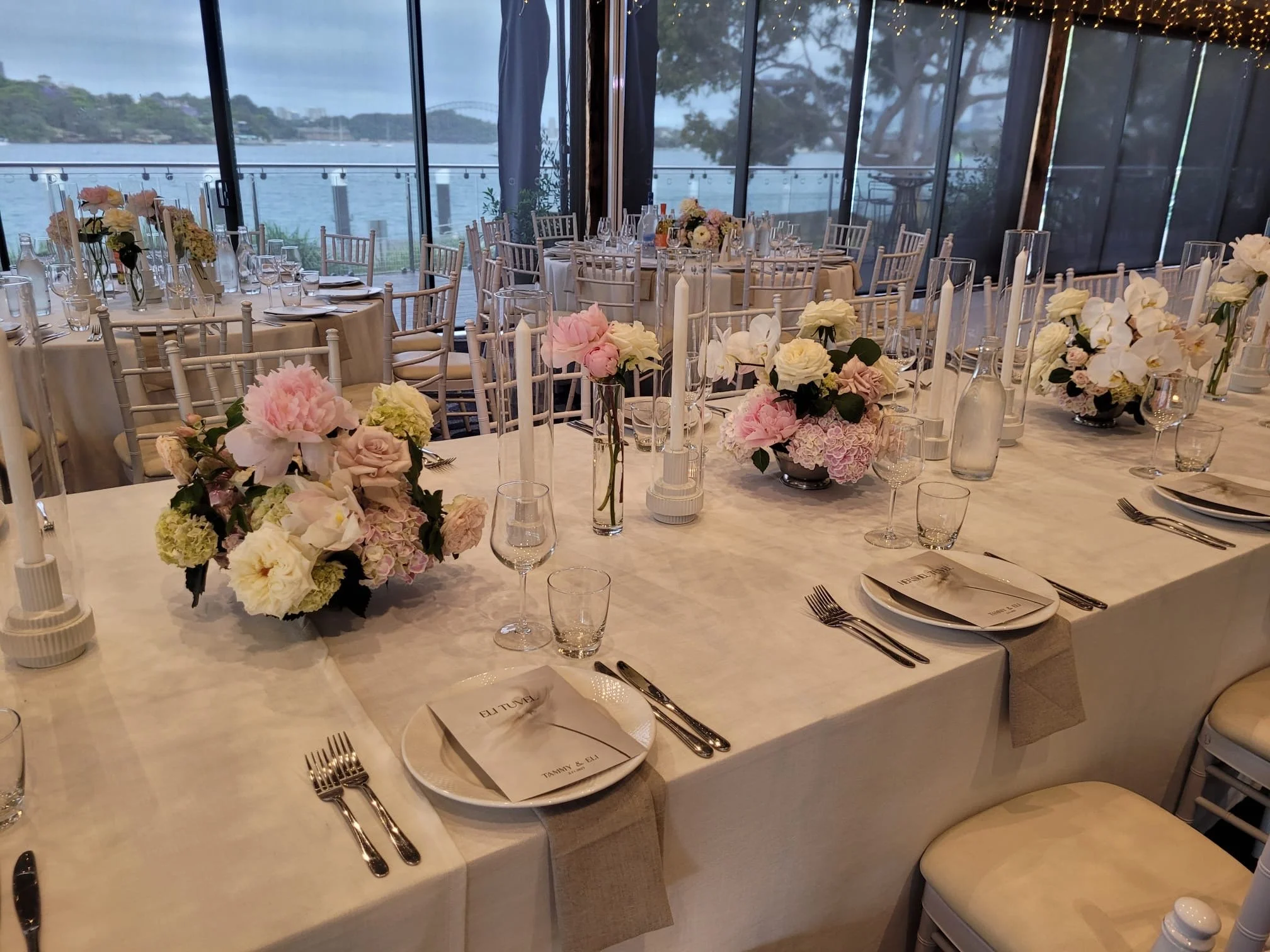 A decorated banquet table set for a formal event with floral centerpieces and a view of water through large windows.