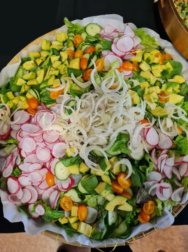 Assorted fresh vegetable salad with radishes, cherry tomatoes, cucumbers, avocados, and onion slices in a basket lined with paper.