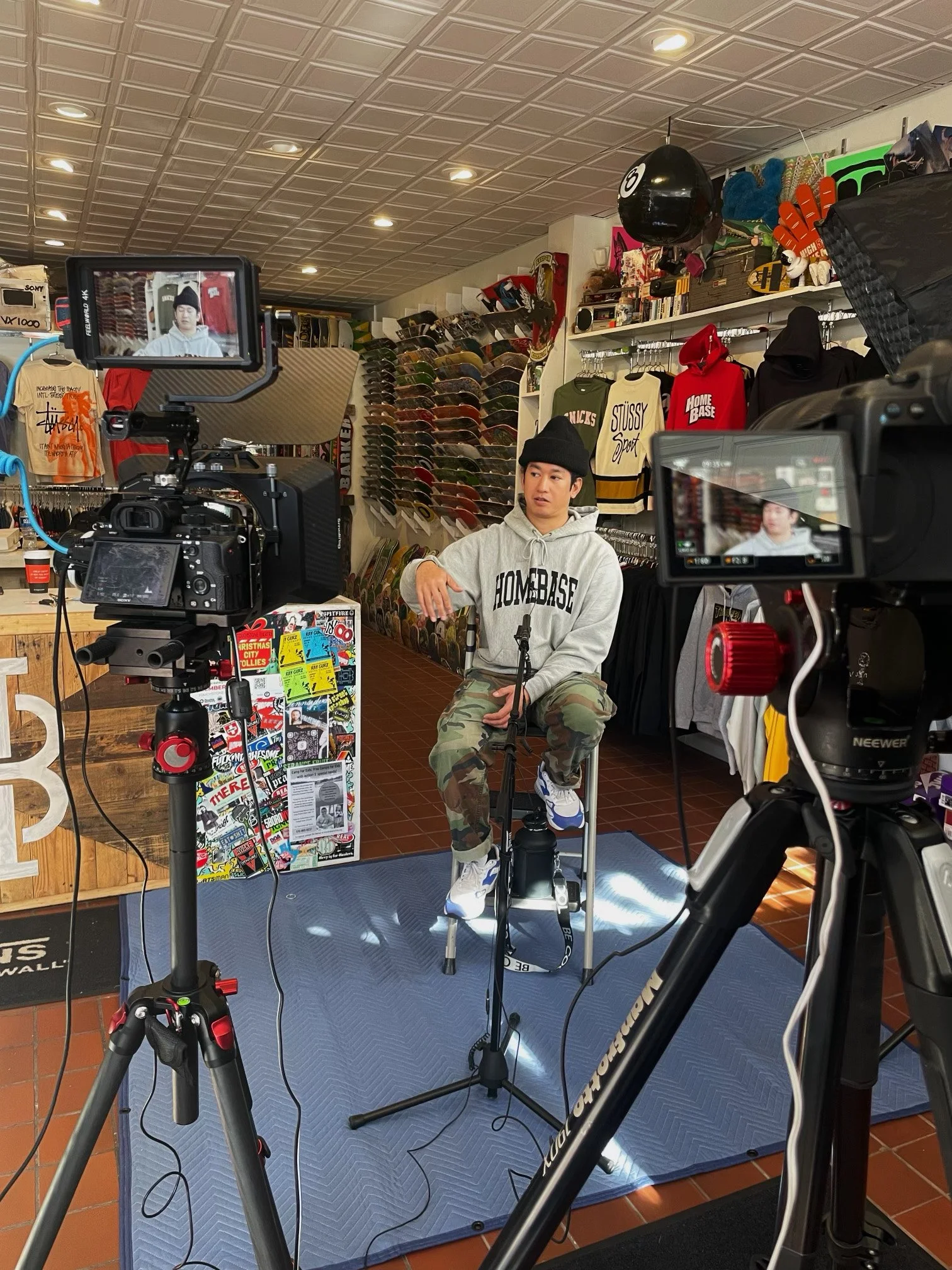 A person in a gray hoodie and camouflage pants is seated in shop, with cameras set up around them recording the scene. The shop has skateboard decks on display on the wall and various clothing items like hoodies and t-shirts hanging on racks.