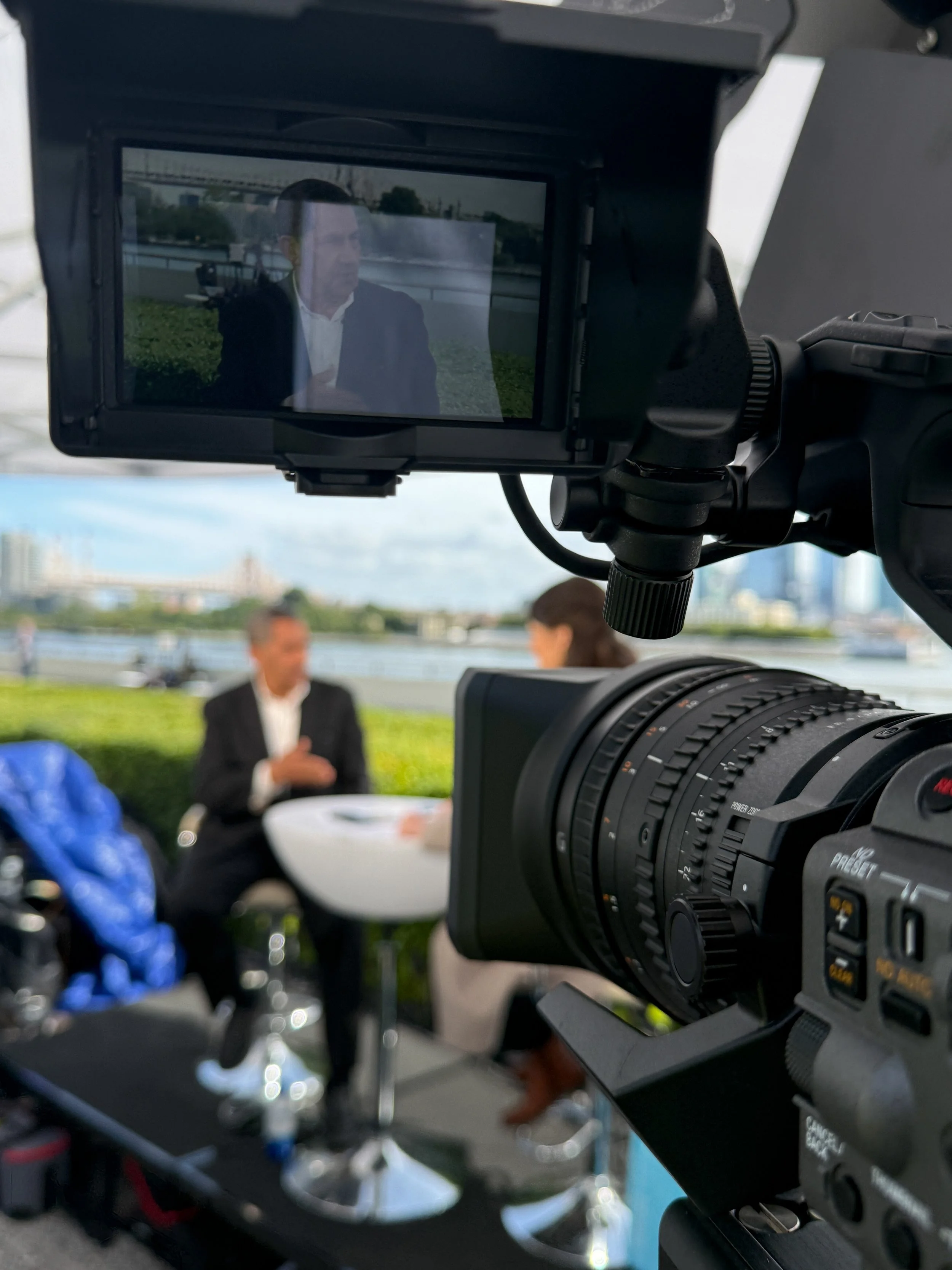 A professional video camera filming an interview with a man in a suit and white shirt, seated outdoors near a river with a city skyline in the background.