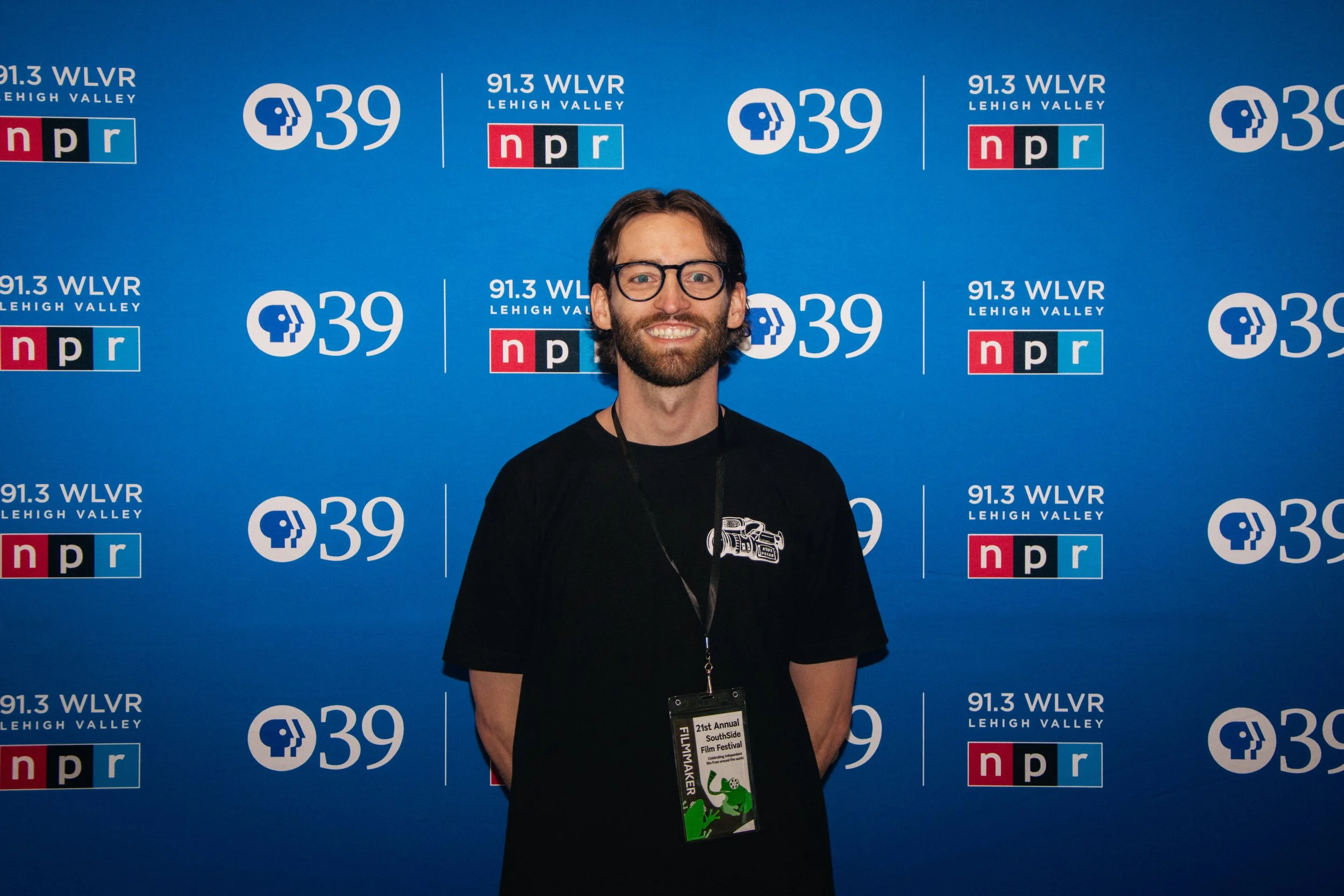 A smiling man with glasses and a beard standing in front of a blue backdrop with logos of 91.3 WLVN Lehigh Valley and NPR, wearing a black T-shirt and a lanyard with a festival badge.