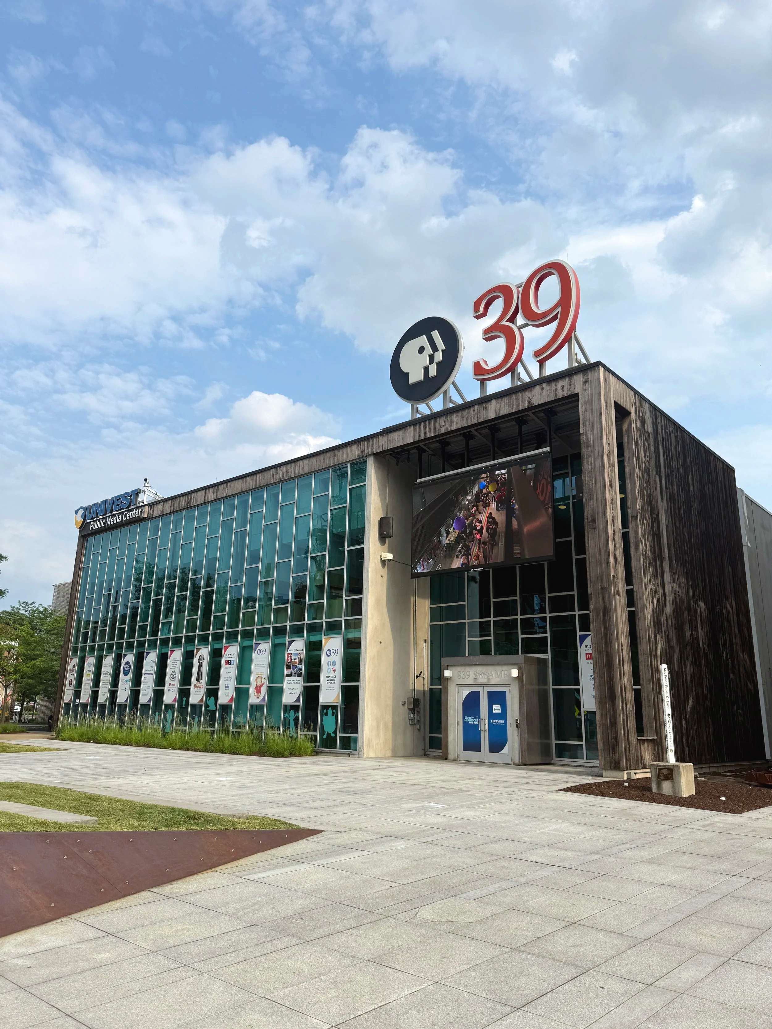 Exterior view of a modern office building with large glass windows, a sign for PBS 39 on the roof, and the number 39 displayed on top. There is a sidewalk and some greenery in front of the building, with a partly cloudy sky above.
