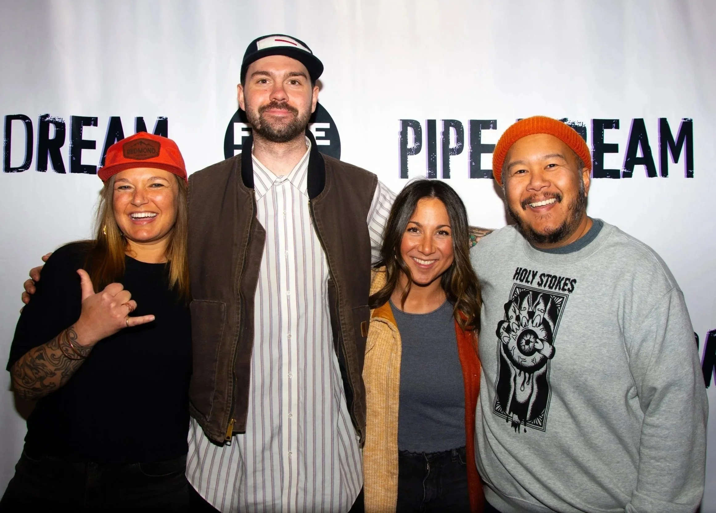 Group of five smiling people standing in front of a white backdrop with words 'DREAM FREE PACE' printed on it. Two women and three men, some with tattoos, wearing casual clothing and colorful hats.