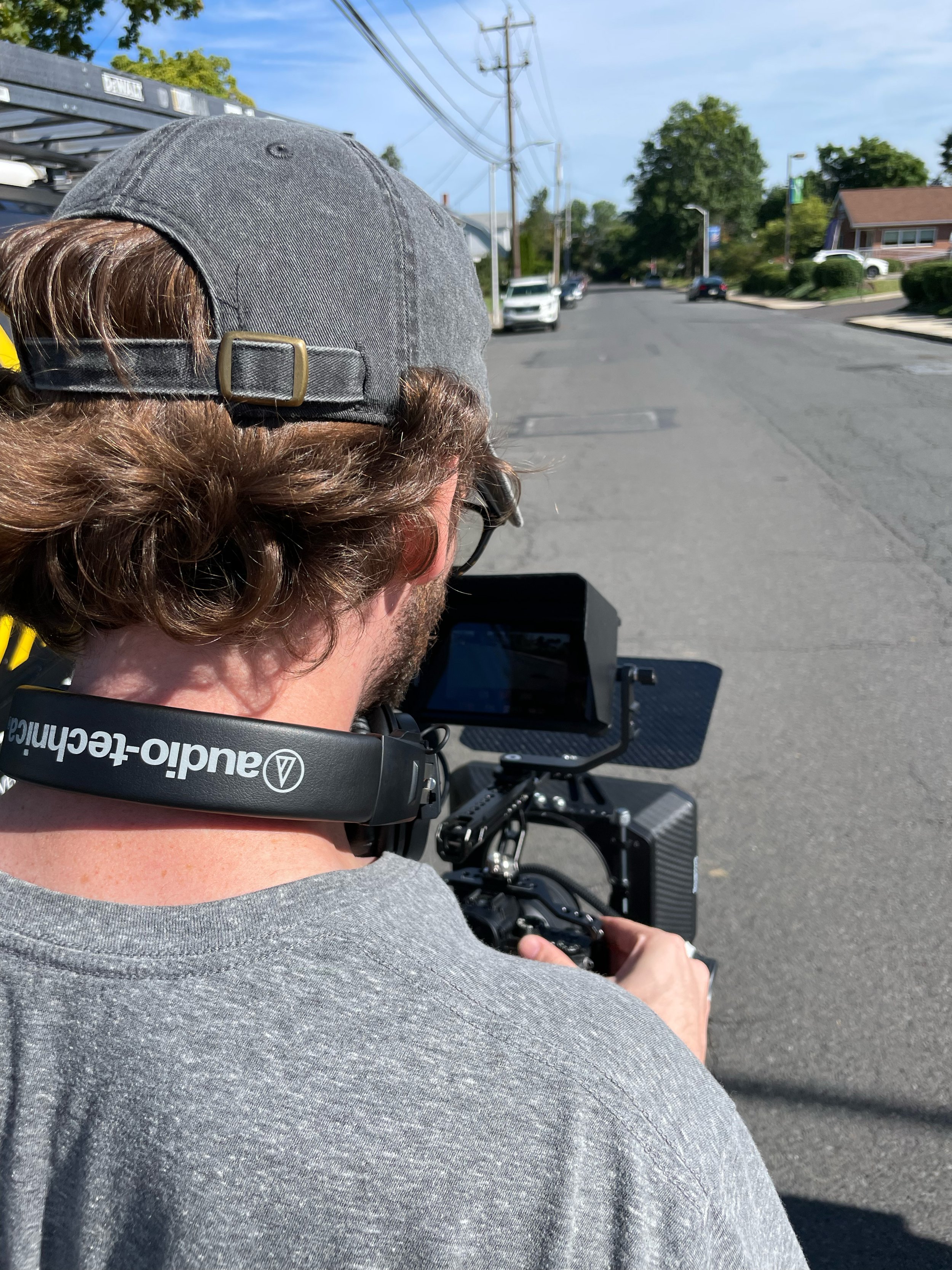 A person wearing a gray baseball cap and glasses, operating a drone with a professional camera in an outdoor street setting with cars, trees, and houses in the background.